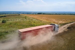 Aerial view of cargo truck driving on dirt road between agricultural wheat fields making lot of dust. Transportation of grain after being harvested by combine harvester during harvesting season.